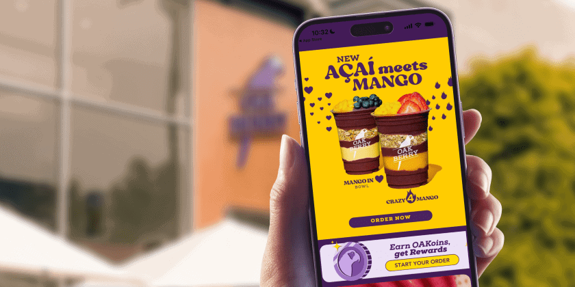Person holding a smartphone showing a promotional Oakberry app screen with the text “NEW AÇAÍ meets MANGO – MANGO IN, BOWL IT – CRAZY 4 MANGO.” The screen displays two açaí bowls with mango and blueberries on a bright yellow background. The Oakberry logo is visible on the building in the blurred background.