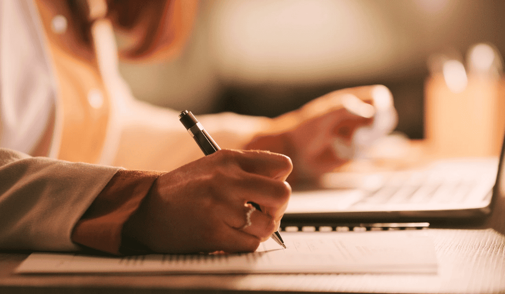 A side view of a hand writing on paper, and the other hand resting on a laptop