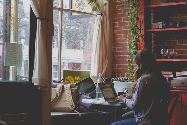 A woman sitting in front of a window at a cafe, with a laptop on the table in front of her. She is typing.