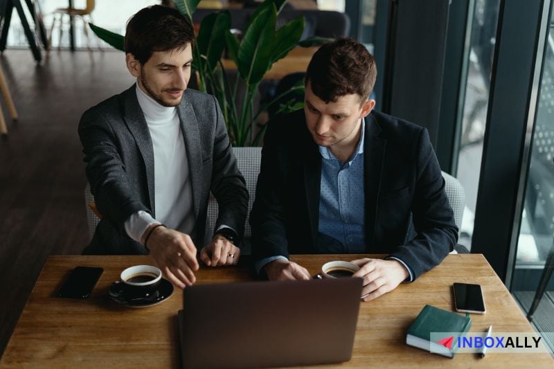 Dois homens em trajes de negócios sentam-se em uma mesa de madeira, discutindo sobre as melhores estratégias de divulgação no LinkedIn enquanto olham para um laptop.  