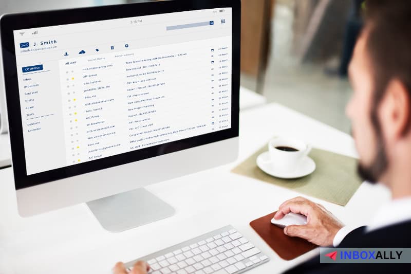 Um homem de terno está trabalhando em um iMac que exibe uma caixa de entrada de e-mail, possivelmente lendo um guia completo para navegar pelas listas negras UCEPROTECTL2. Uma xícara de café está sobre a mesa ao lado dele.