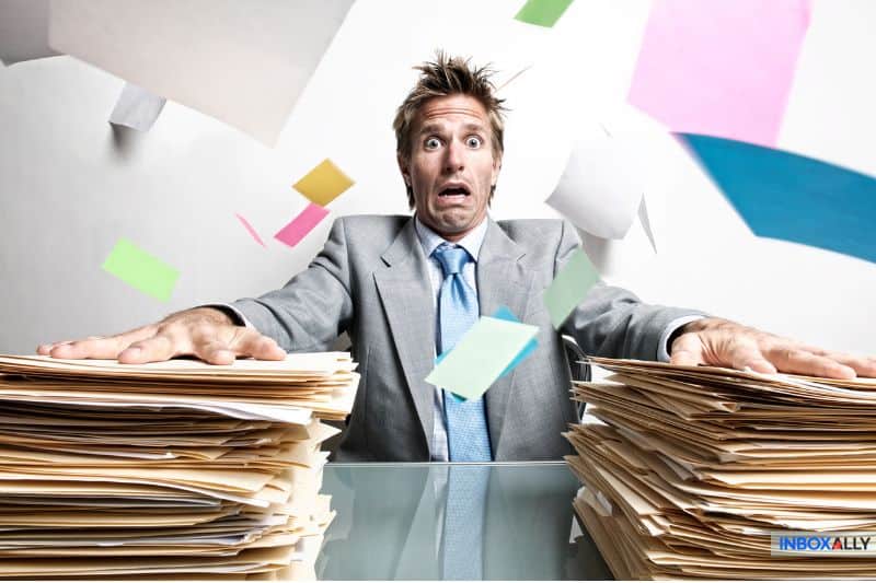 Um homem de terno parece sobrecarregado enquanto papéis coloridos voam ao seu redor, cercado por duas grandes pilhas de arquivos em uma mesa, lembrando sua caixa de entrada caótica antes da sincronização de e-mails simplificar sua vida.