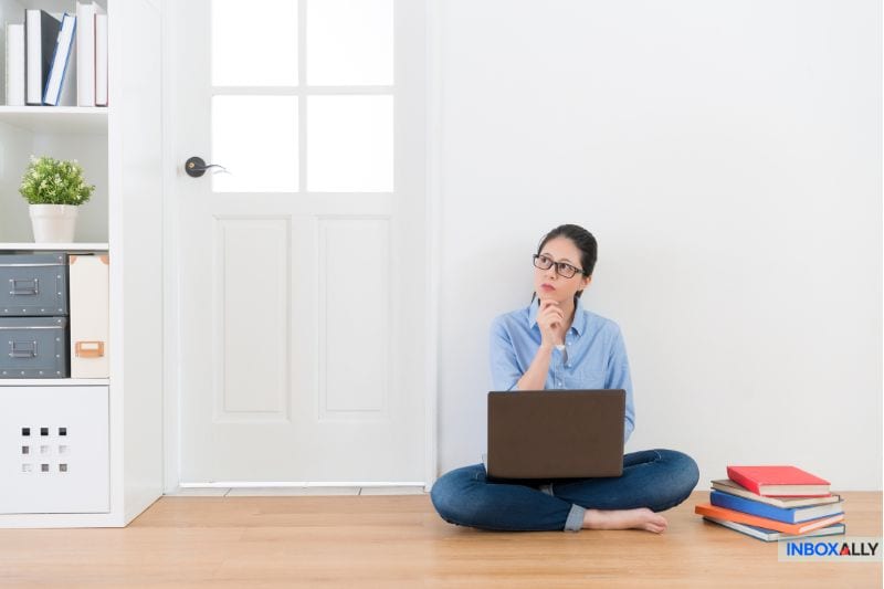 A woman sits thoughtfully on the floor with her laptop, exploring Top Picks for email solutions. Books are stacked beside her as she leans against the white wall in front of a door, delving into discussions about the Best Sendgrid options out there.
