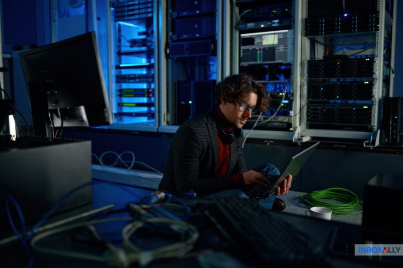 Uma pessoa está sentada no chão de uma sala de servidores trabalhando em um laptop, configurando um SMTP Relay entre cabos de rede e racks de servidores.