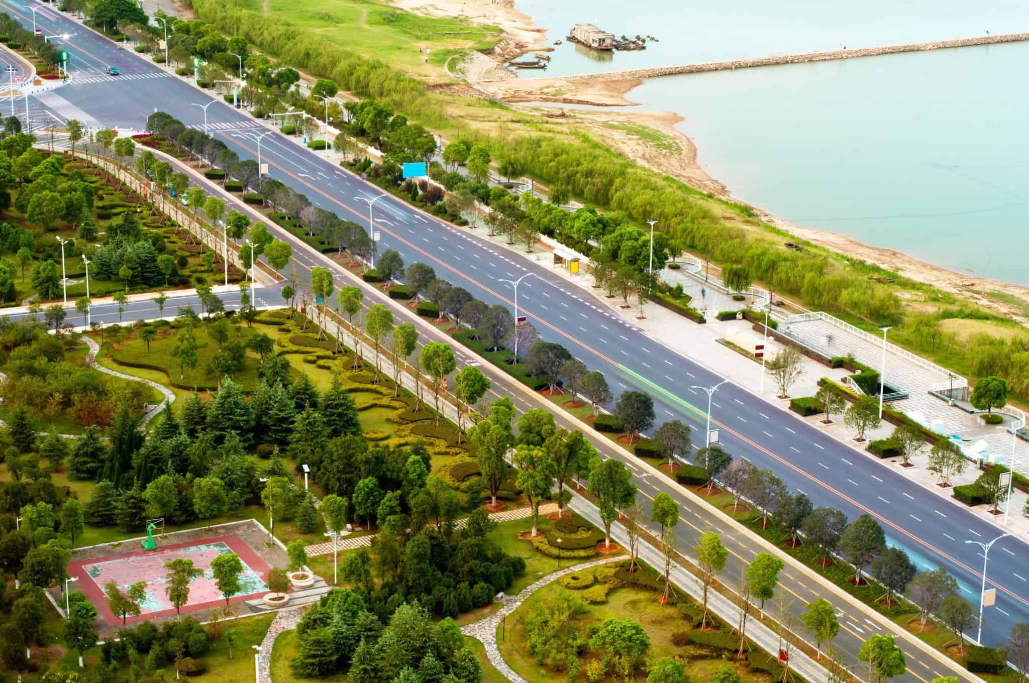 Aerial view of a coastal park with winding paths, manicured lawns, and a diagonal road beside a calm shoreline.