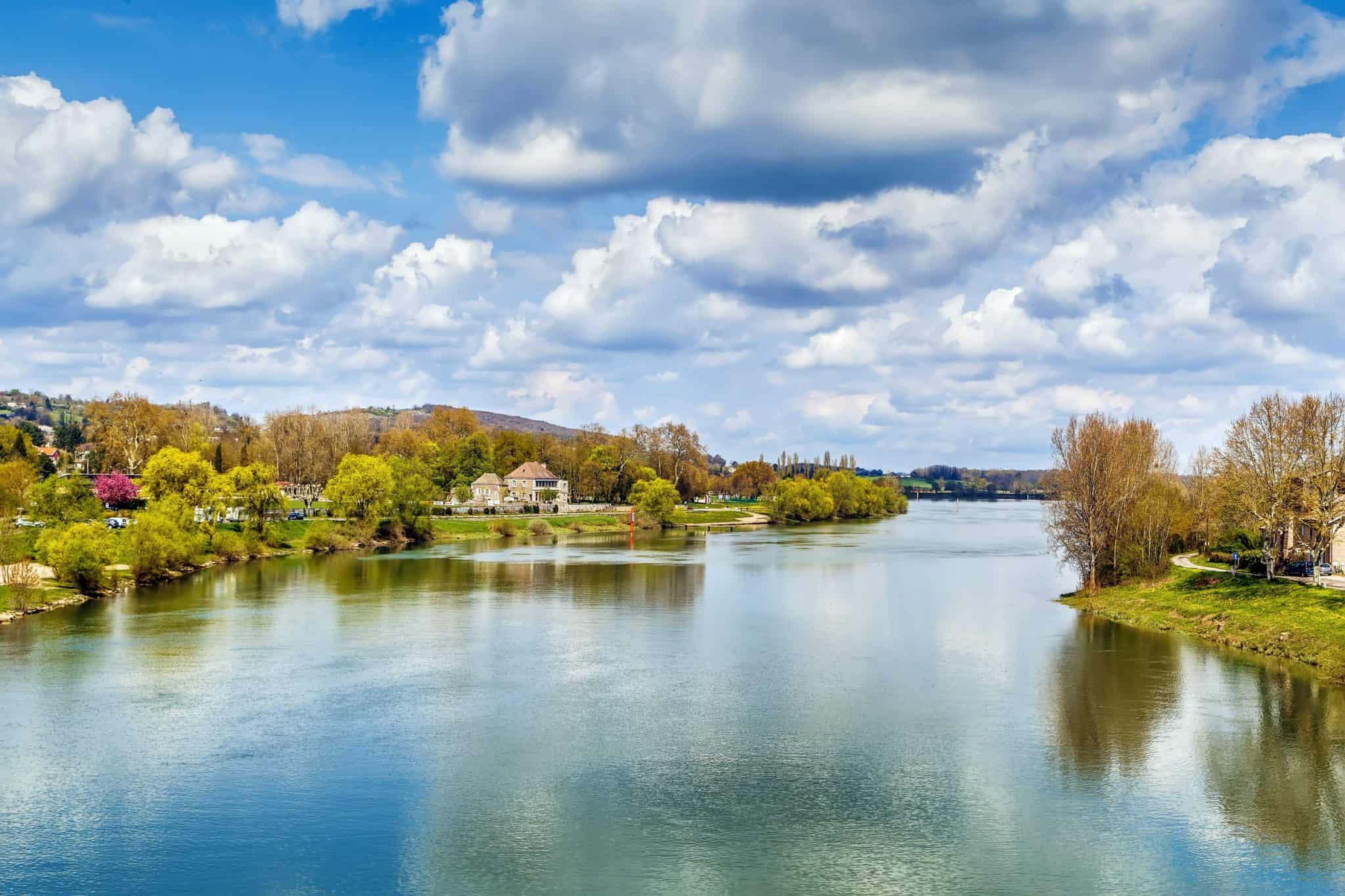 Riverside scene with calm water, trees in spring foliage, and houses lining the riverbank under a partly cloudy blue sky.