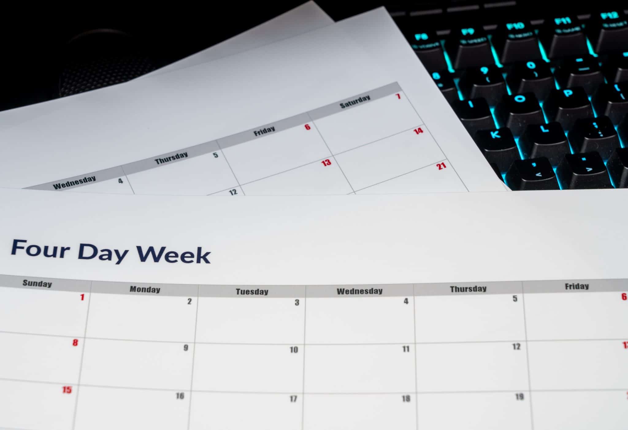 Close-up of a printed monthly calendar page with a four-day-week header, calendar grid and red dates, with a backlit keyboard in the background