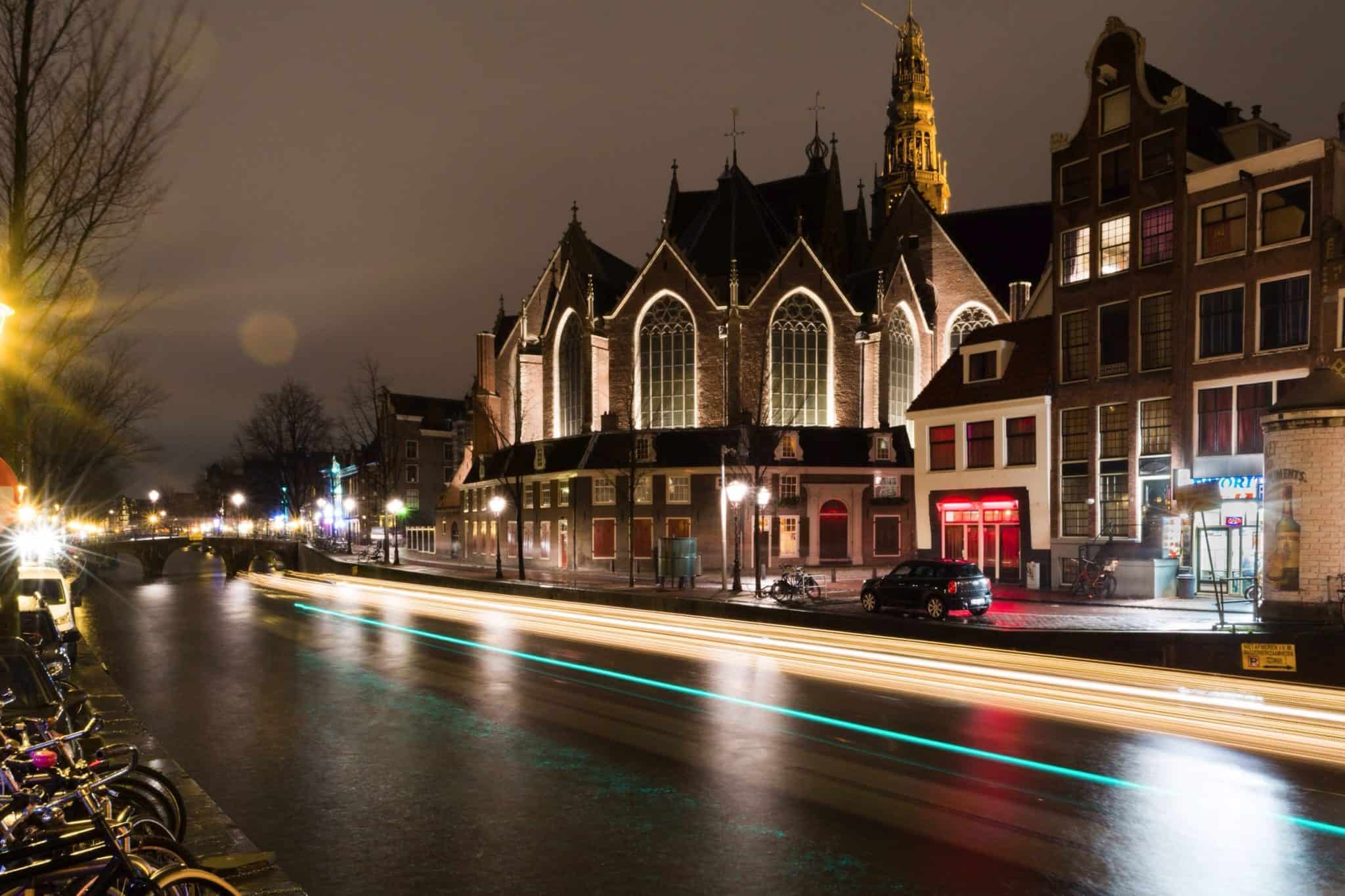 Night scene of a historic Gothic church on a canal street with light trails from passing cars and a row of buildings beside the water.