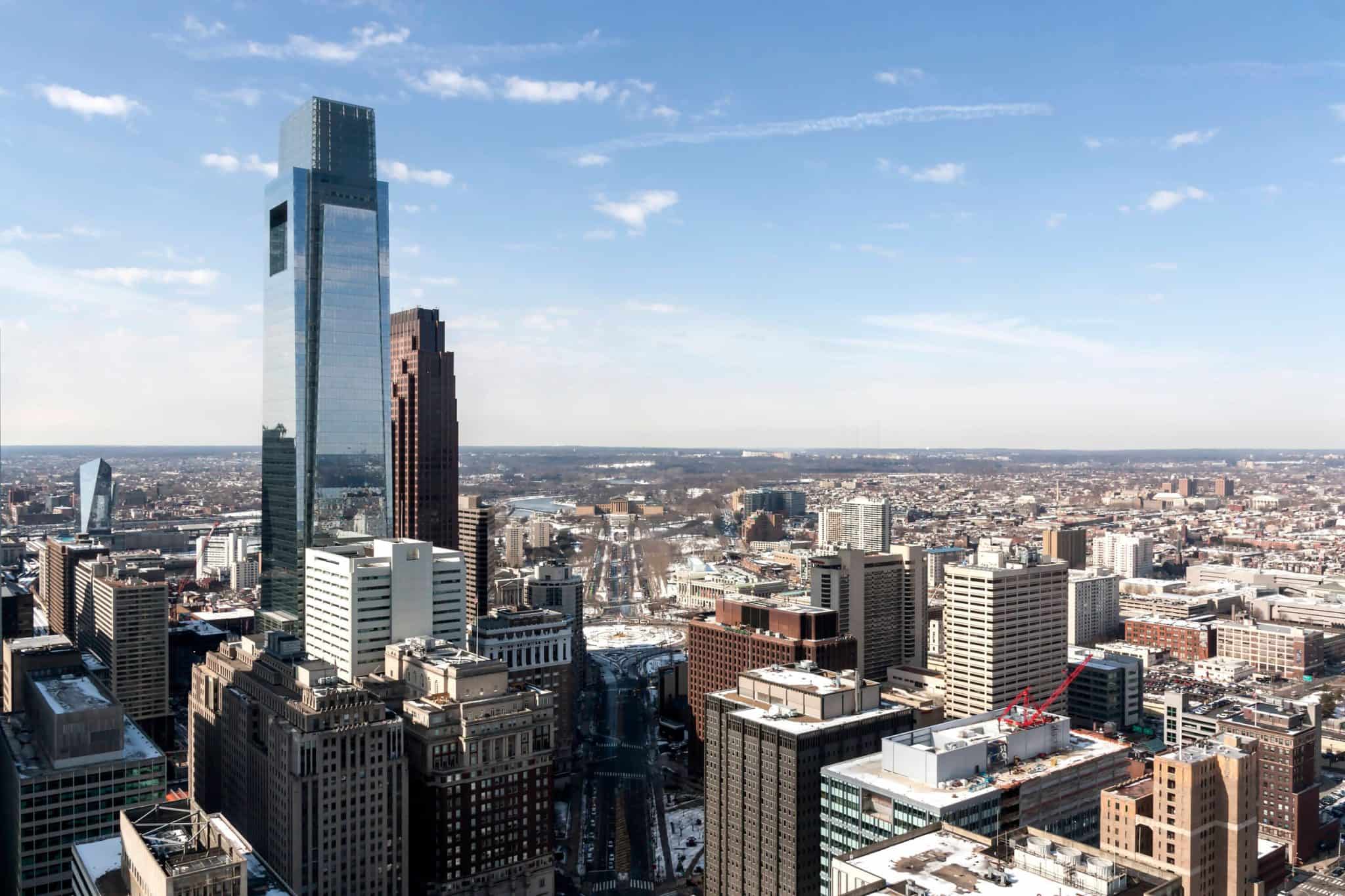 Tall glass skyscraper rises among a dense downtown skyline under a clear blue sky, on a sunny day close to noon.