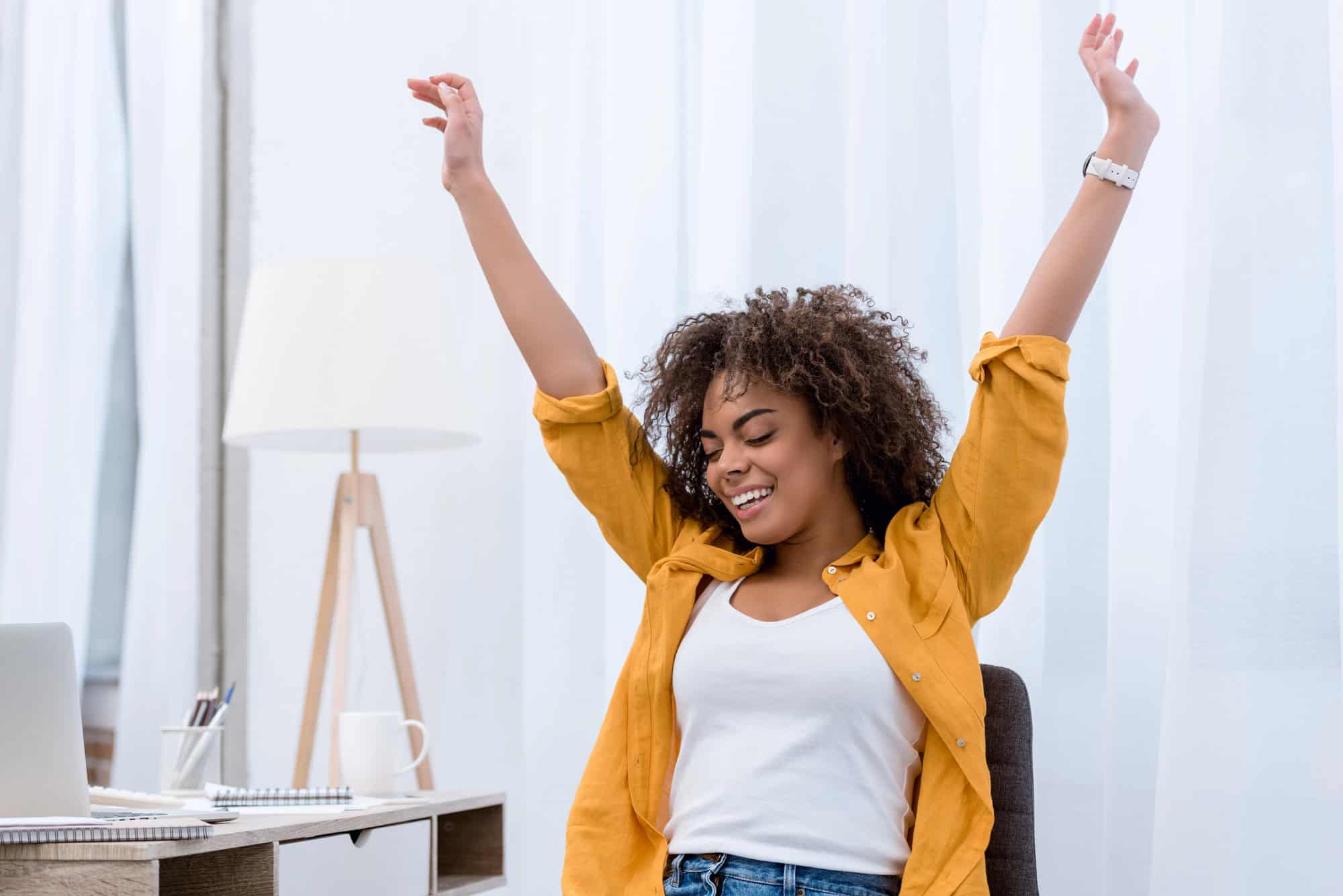 Woman with curly hair in a mustard shirt raising arms in celebration in a bright home office setting with a laptop nearby.