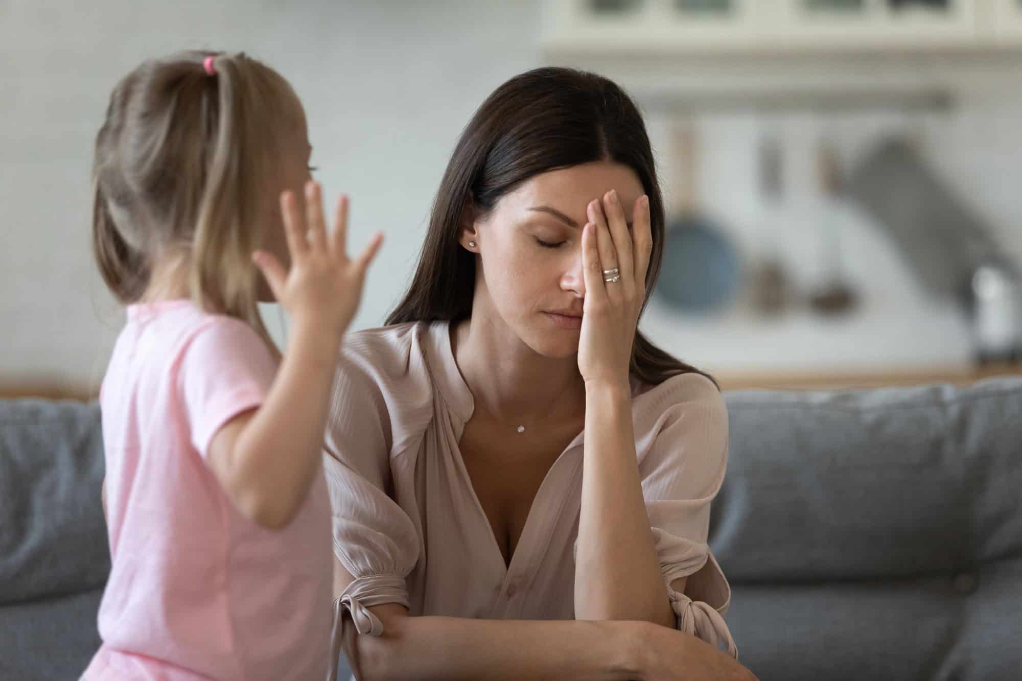 Woman sitting on a couch with her hand covering her face, looking stressed as a young girl reaches toward her in a pink shirt.