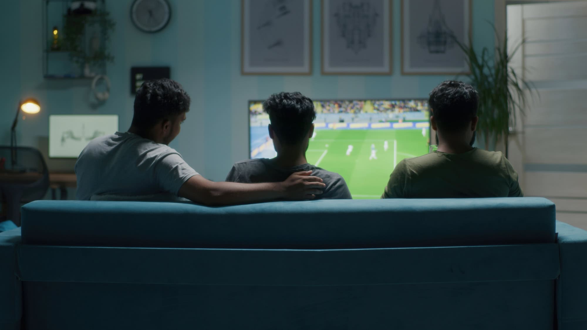 Three friends sit on a blue sofa watching a soccer match on a television in a cozy living room.