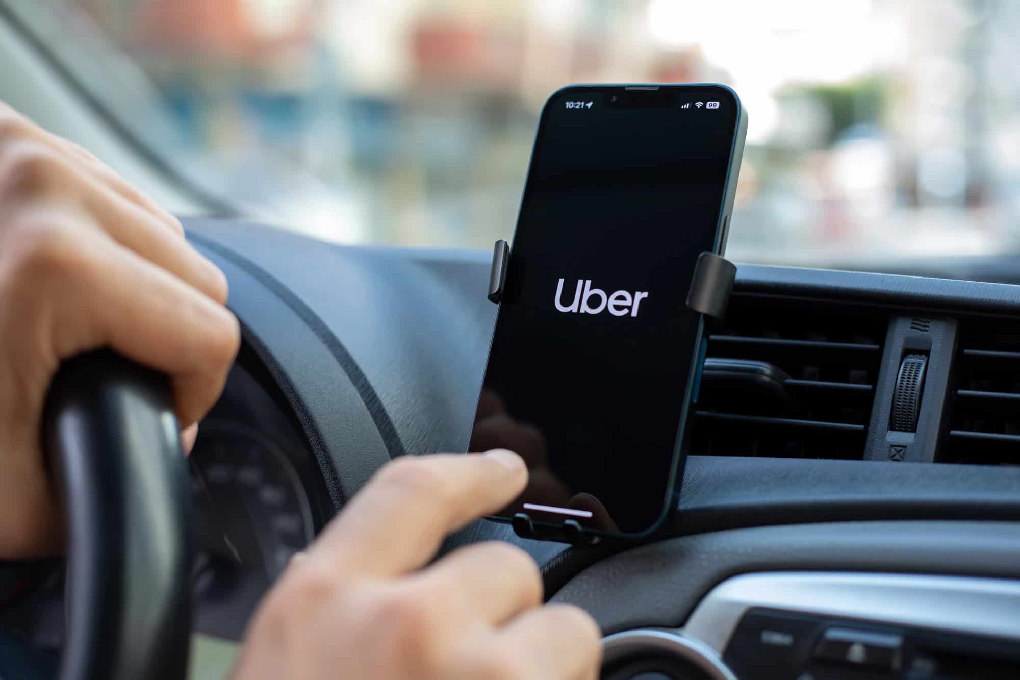 Driver's hands on the steering wheel with a smartphone displaying the Uber app in a dashboard mount.