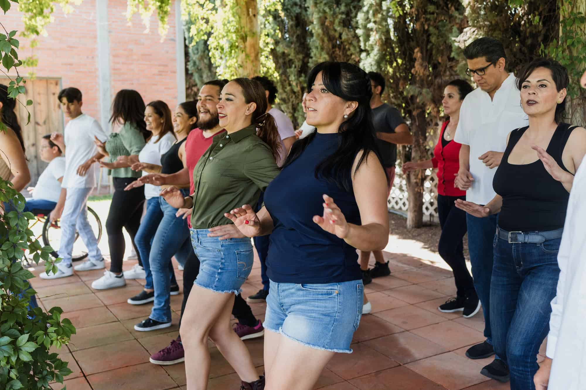 Diverse group of adults dancing in an outdoor courtyard, smiling and moving together among trees and brick walls.