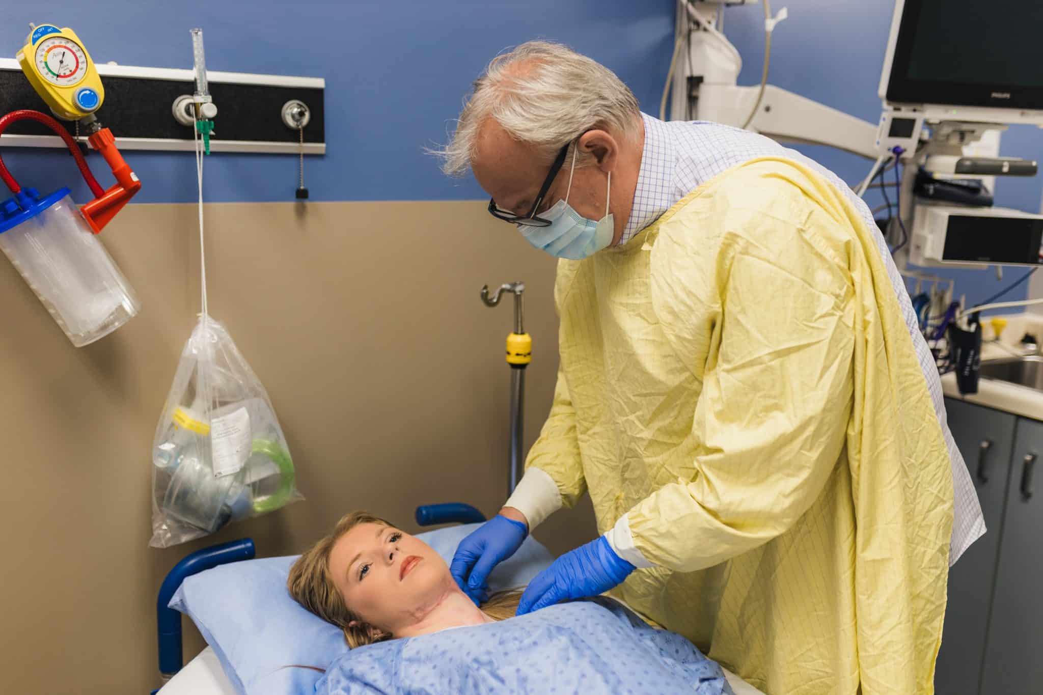 Hospital scene: clinician in a yellow gown and mask tending to a patient lying in bed.