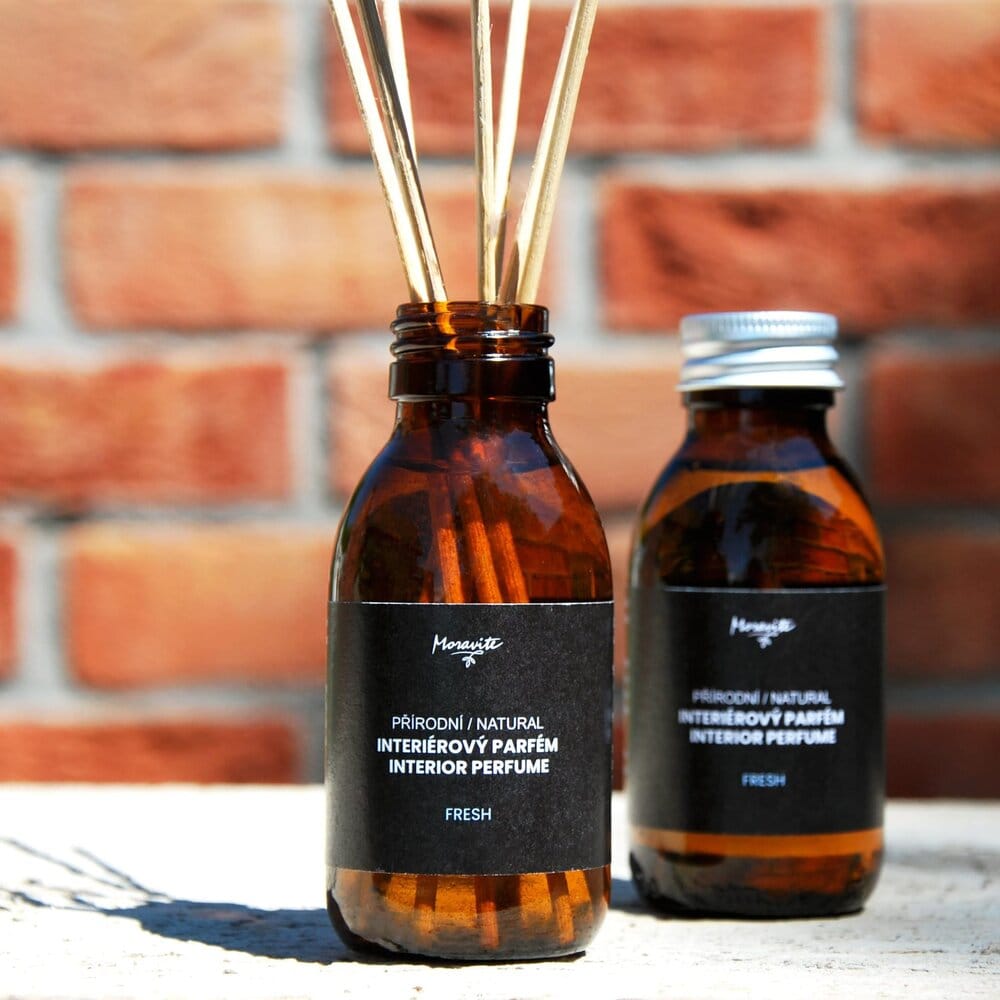 Dvě tmavé skleněné lahvičky s černou etiketou. Na etiketě je bílé logo Moravité a bílé nápisy PŘÍRODNÍ a NATURAL INTERIÉROVÝ PARFÉM INTERIOR PERFUME FRESH. Levá lahvička je otevřená s vloženými dřevěnými tyčinkami, pravá je uzavřená stříbrným víčkem. Pozadí tvoří cihlová zeď.