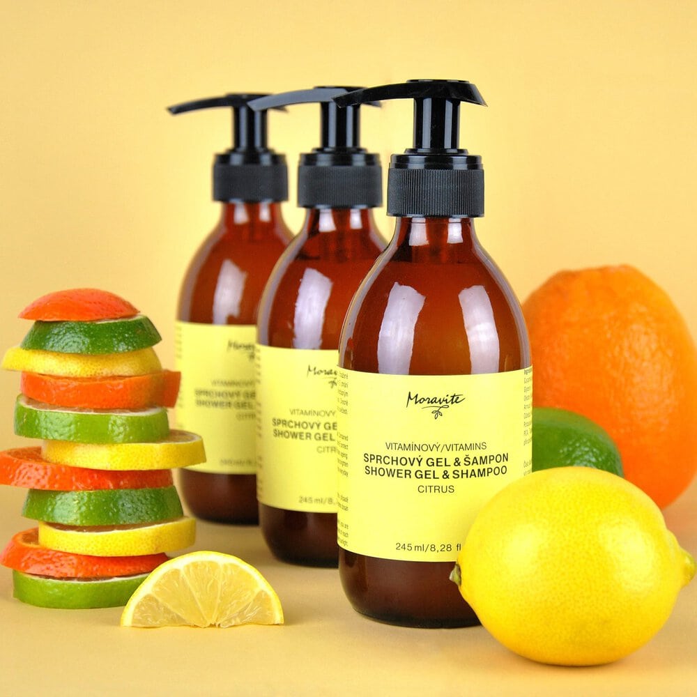 Tři lahvičky Vitamínového sprchového gelu a šamponu Moravité Citrus (245 ml) obklopené čerstvými citrony, limetkami a pomeranči.
