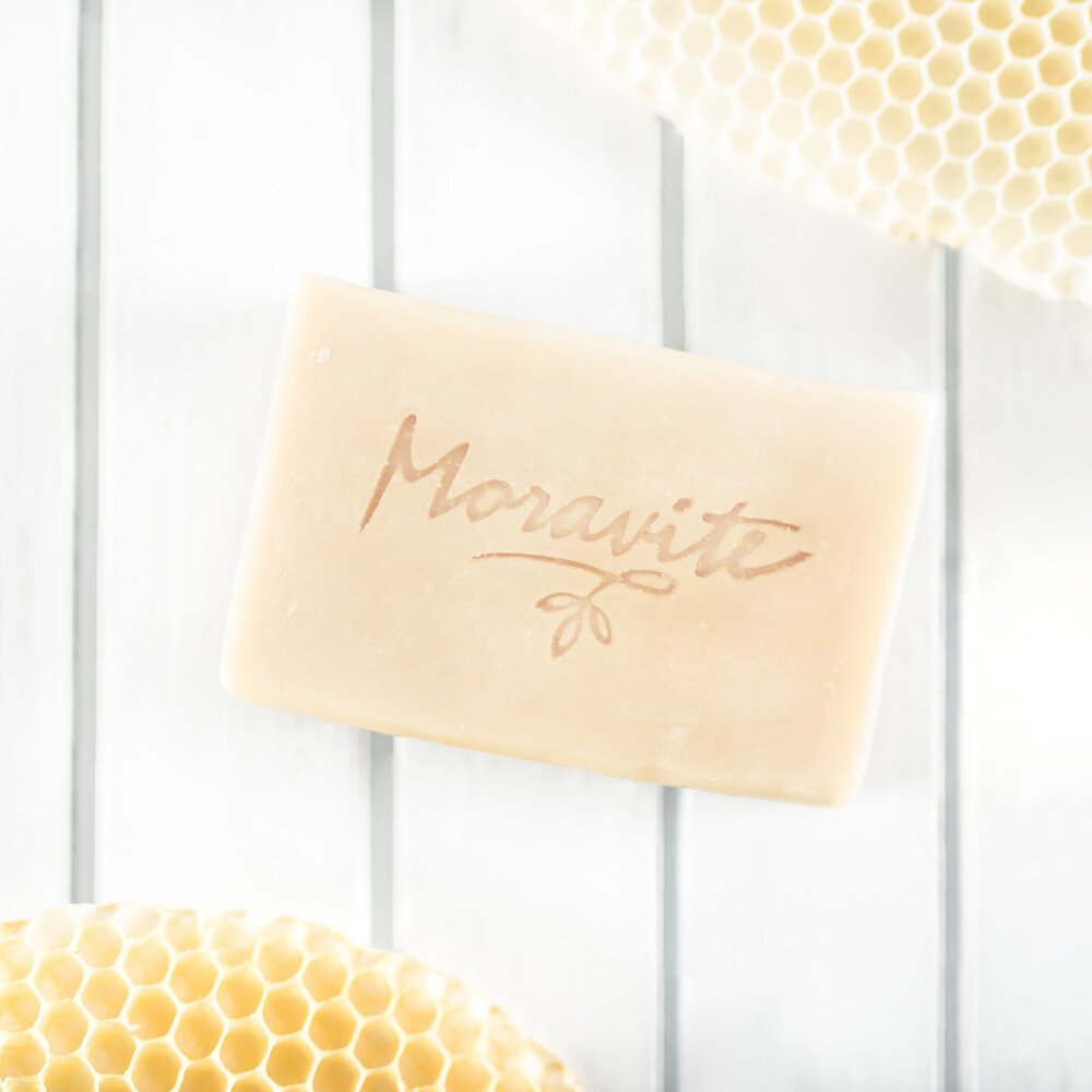 Světlé přírodní mýdlo s vyraženým logem Moravité na bílém mřížkovaném pozadí s detaily včelích pláství.