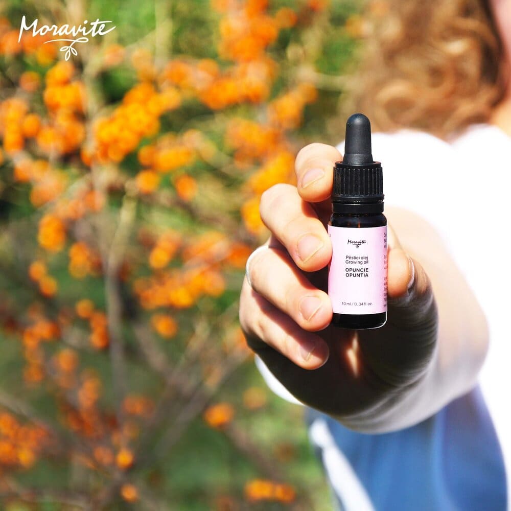 Ruka držící lahvičku pěsticího oleje Moravité Opuncie (10 ml) na pozadí rozostřeného oranžového keře rakytníku.
