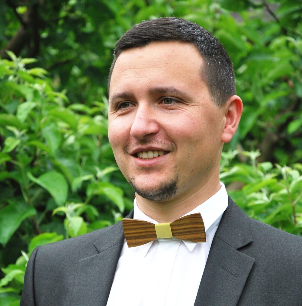 Portrét muže v tmavém obleku s pruhovaným dřevěným motýlkem se světlým středem na zeleném pozadí.