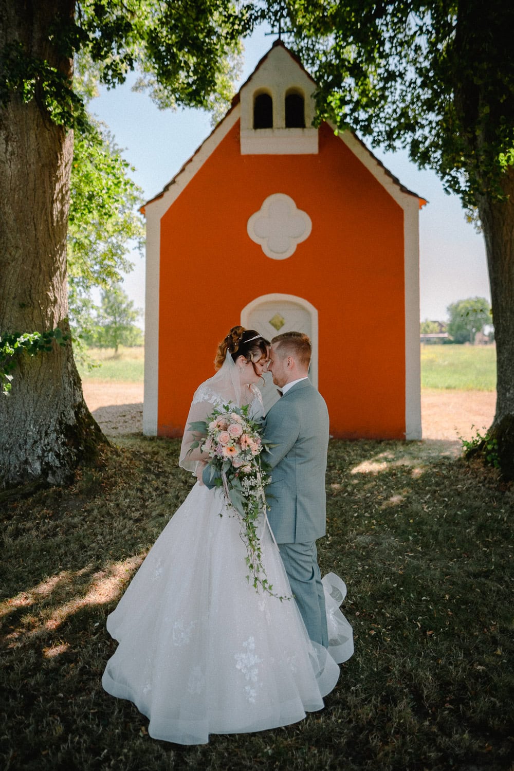 Braut und Bräutigam verbringen einen Moment vor einer kleinen, orangefarbenen Kapelle, die von Bäumen umgeben ist. Die Braut trägt ein weißes Kleid und hält einen Blumenstrauß, während der Bräutigam einen hellgrauen Anzug trägt. Sonnenlicht fällt durch die Blätter und wirft gesprenkelte Schatten – eine perfekte Aufnahme von ihrem Hochzeitsfotografen Kallmünz.