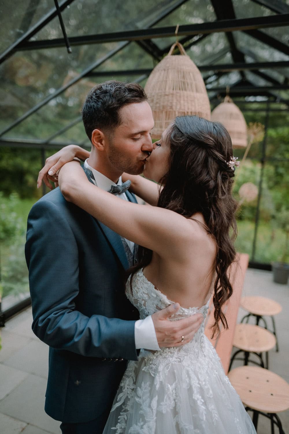 Bei Hochzeit Schloss Pirkensee küssen sich Braut und Bräutigam unter dem Glasdach. Das Spitzenbrautkleid der Braut leuchtet sanft, während der Bräutigam in dunklem Anzug und Fliege neben ihr steht. Holzlaternen hängen anmutig über ihnen und werfen sanfte Schatten auf den Holztisch im Hintergrund.