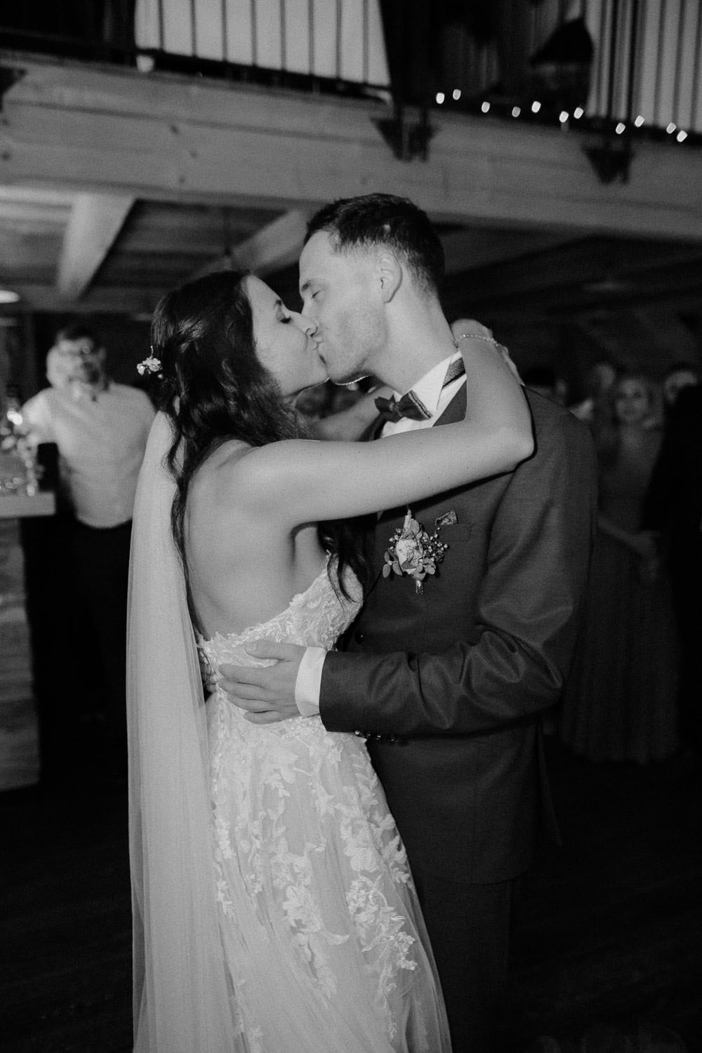 Bei ihrer Hochzeit im Schloss Pirkensee küssen sich Braut und Bräutigam während des Empfangs zärtlich. Die Braut trägt ein schulterfreies Kleid mit langem Schleier und der Bräutigam einen Anzug mit Ansteckblume. Sie umarmen sich auf der schwach beleuchteten Tanzfläche, während im Hintergrund die Gäste fröhlich feiern.