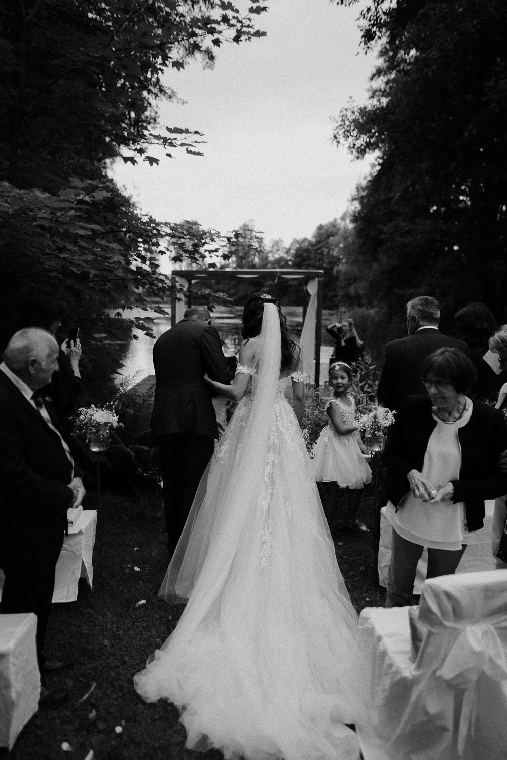 Eine Braut in einem langen Kleid und Schleier geht Arm in Arm mit einem älteren Mann auf einen Bräutigam zu bei einer Zeremonie im Freien auf Schloss Pirkensee. Gäste stehen auf beiden Seiten. Die Umgebung ist üppig mit Bäumen und einem nahe gelegenen Gewässer, wunderschön in Schwarzweiß festgehalten.