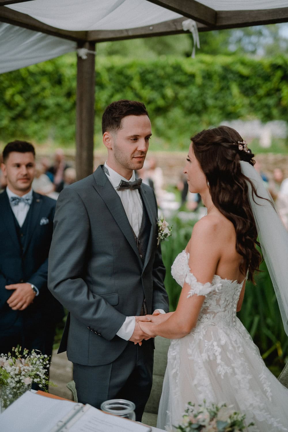 Braut und Bräutigam stehen sich während ihrer Hochzeitszeremonie im bezaubernden Schloss Pirkensee unter einem Baldachin gegenüber und halten Händchen. Der Bräutigam trägt einen grauen Anzug mit Fliege und die Braut ein weißes Spitzenkleid mit Schleier. Im Hintergrund sind einige Gäste zu sehen.