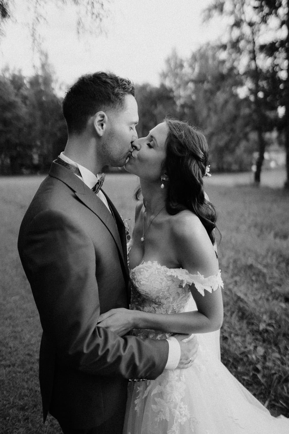 In einem zeitlosen Schwarz-Weiß-Moment auf Schloss Pirkensee küssen sich die Braut in einem schulterfreien Spitzenkleid und der Bräutigam in einem eleganten Anzug zärtlich, umgeben von Grasfeldern und hoch aufragenden Bäumen.