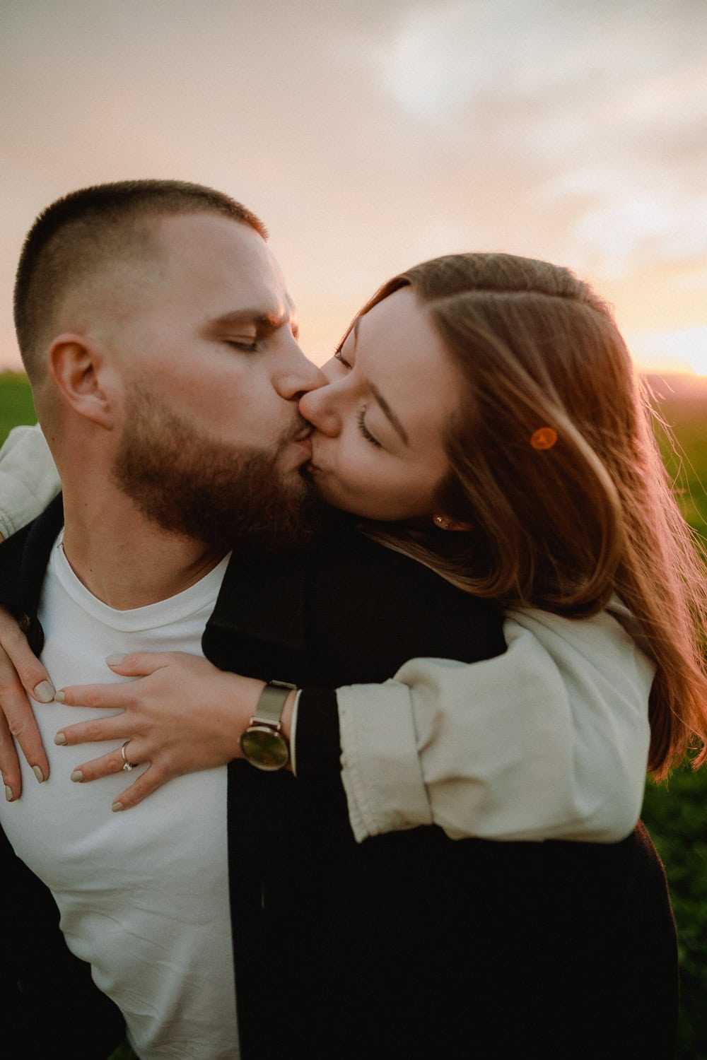 Während ihres Paarshootings küsst sich ein Paar bei Sonnenuntergang auf einer Wiese zärtlich. Der Mann hat einen Bart und trägt eine schwarze Jacke, während die Frau in ihrem beigen Mantel ihre Arme um seine Schultern legt. Der Himmel ist sanft in warmen Farbtönen beleuchtet und fängt die Essenz ihrer Verlobung ein.