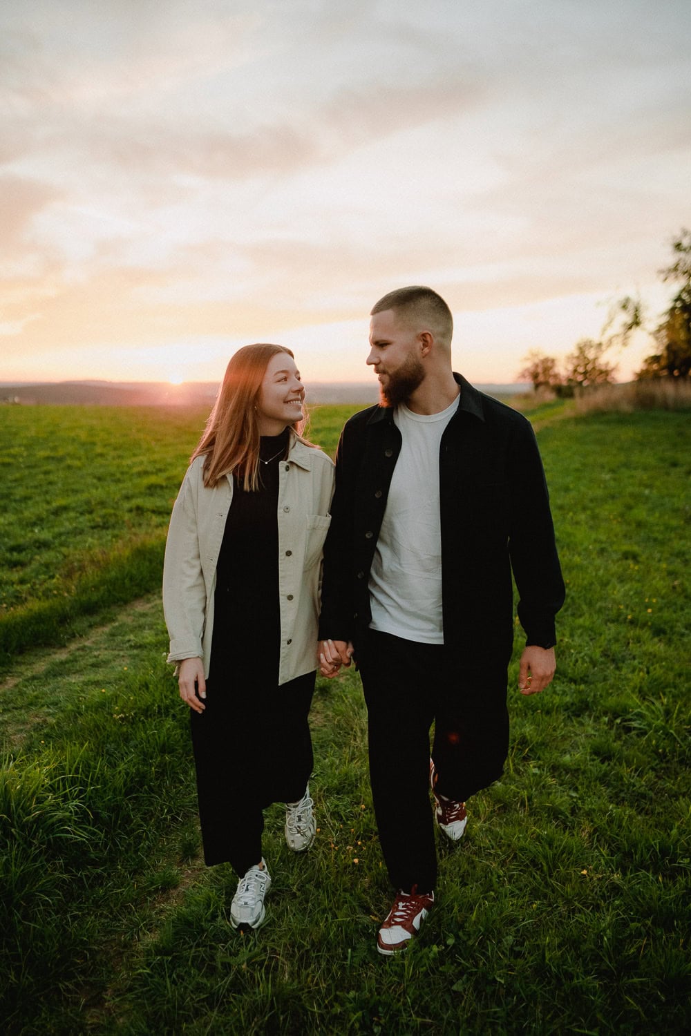 Ein Paar genießt ein romantisches Paarshooting, während sie bei Sonnenuntergang Hand in Hand über eine Wiese laufen. Die Frau trägt eine helle Jacke und Turnschuhe, während der Mann eine dunkle Jacke und rote Schuhe trägt. Beide lächeln und schauen sich an, ihre Liebe leuchtet unter dem orangefarbenen und rosa Himmel.
