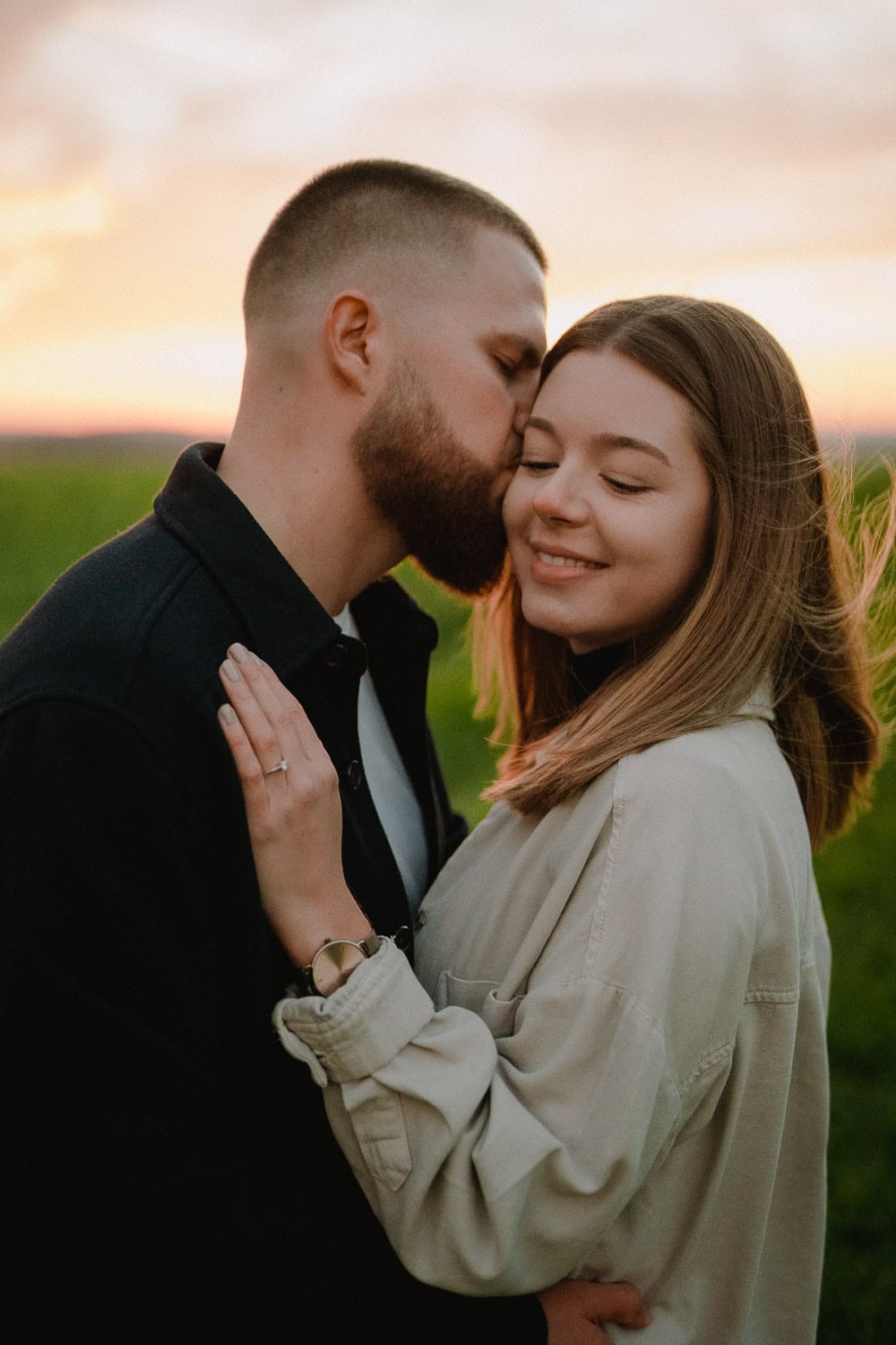 Bei Sonnenuntergang, während ihres Paarshootings, küsst ein bärtiger Mann seine lächelnde Verlobte auf einem Feld auf die Stirn. Der warme Glanz umhüllt sie, während sie eine helle Jacke trägt und er eine dunkle, was die Essenz ihrer Verlobung einfängt.
