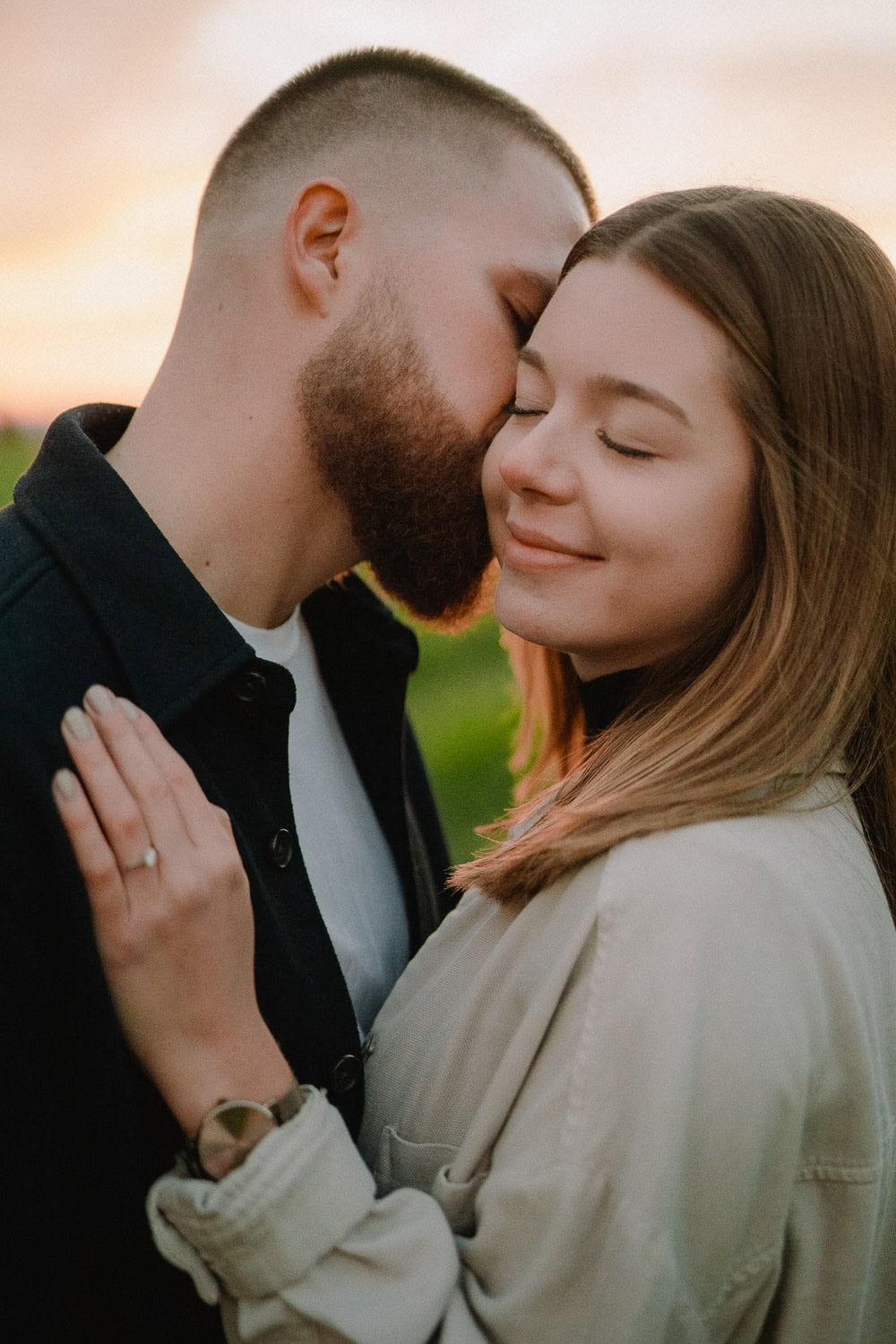 Ein Paar umarmt sich zärtlich auf einem Feld bei Sonnenuntergang während seiner Verlobung. Der Mann beugt sich vor, um die Frau auf die Wange zu küssen, während sie mit geschlossenen Augen lächelt. Ihre Hand ruht auf seiner Schulter und zeigt einen Ring. Beide tragen leichte Jacken und der Himmel leuchtet warm im Hintergrund ihres Paarshootings.