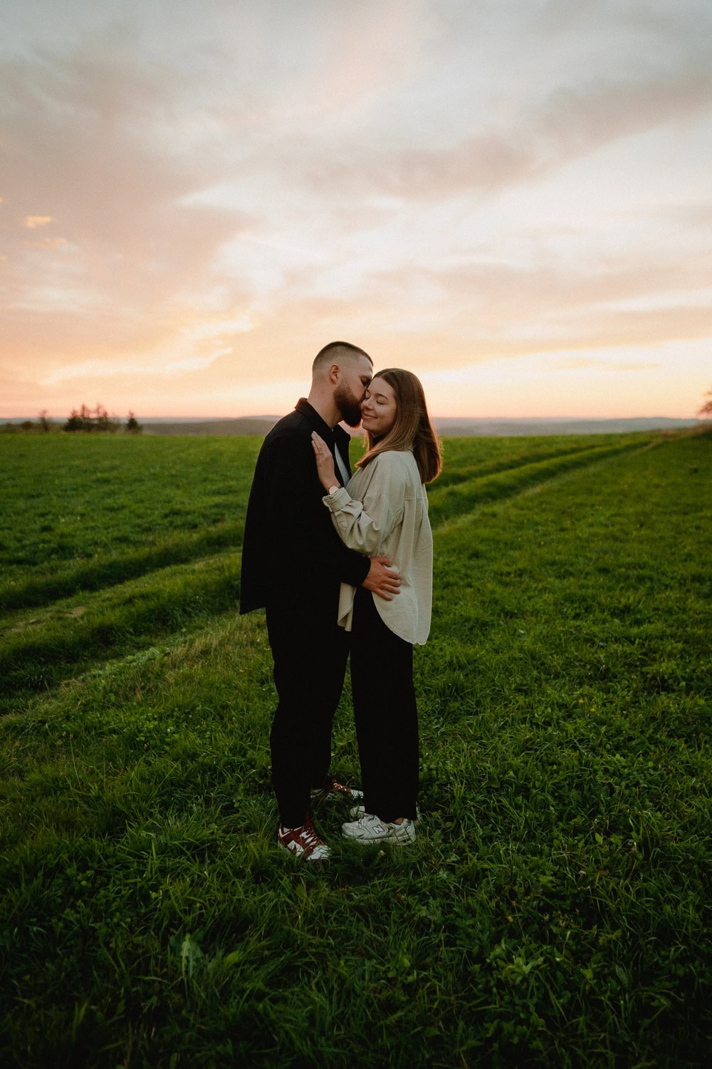 Ein Paar steht bei Sonnenuntergang auf einer Wiese, umarmt sich und blickt sich während seiner Verlobung an. Der Himmel ist in eine Mischung aus Orange- und Rosatönen getaucht und umhüllt sie in einem ruhigen, offenen Raum, der perfekt für ihr intimes Paarshooting ist.
