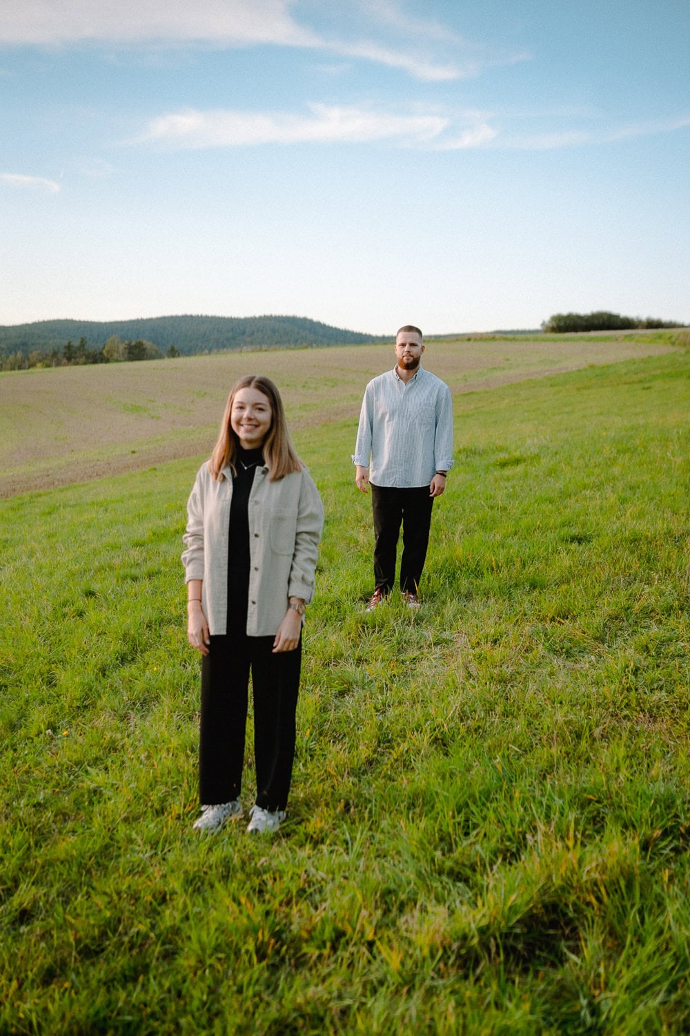 Eine Frau und ein Mann stehen auf einer Wiese unter einem klaren blauen Himmel und fangen die Essenz einer romantischen Verlobung ein. Die Frau steht im Vordergrund und lächelt mit den Händen in den Taschen, während der Mann weiter hinten steht und in die Kamera blickt. Sanfte Hügel bilden die malerische Kulisse ihres Paarshootings.