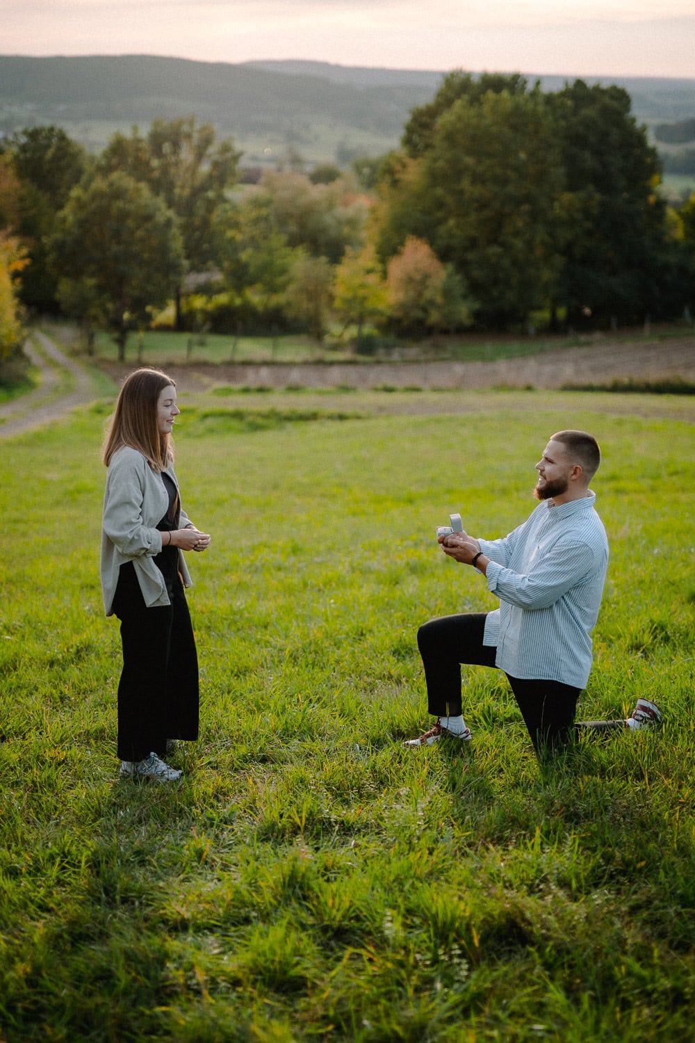 Ein Mann kniet auf einem Knie, hält eine Ringschachtel in der Hand und macht einer Frau auf einer malerischen Wiese einen Heiratsantrag. Dieser bezaubernde Moment der Verlobung entfaltet sich mit Bäumen im Hintergrund unter dem sanften Abendlicht und fängt ein zeitloses Paarshooting ein.