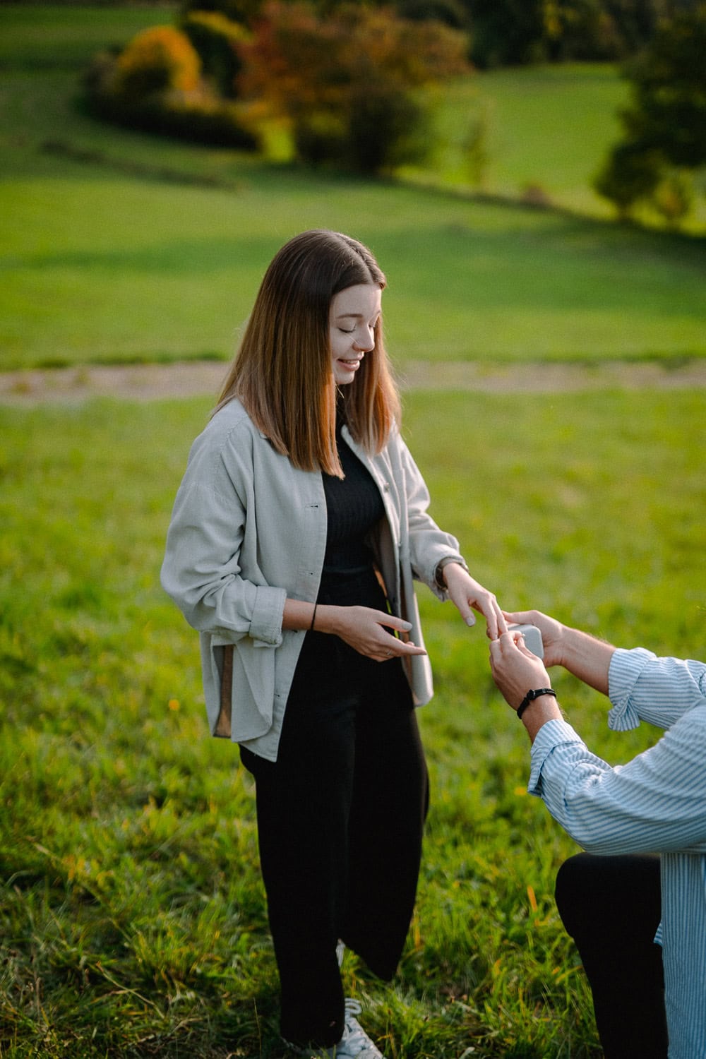 Auf einer ruhigen Wiese mit Bäumen als Zeugen kniet eine Person nieder, um in einem bezaubernden Verlobungsmoment einen Ring anzubieten. Die stehende Person lächelt herzlich und streckt in dieser malerischen Paarshooting-Szene der Liebe und Hingabe ihre Hand aus.
