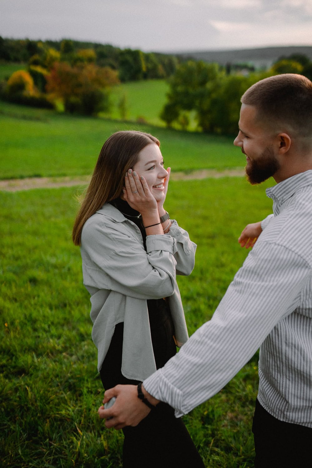 Auf einer Wiese genießt ein Paar einen Verlobungsmoment, lächelt sich an, umgeben von sanften Hügeln und Bäumen. Die Frau berührt ihr Gesicht mit beiden Händen, während der Mann während des Paarshootings nach ihr greift, beide glücklich und entspannt unter einem bewölkten Himmel.