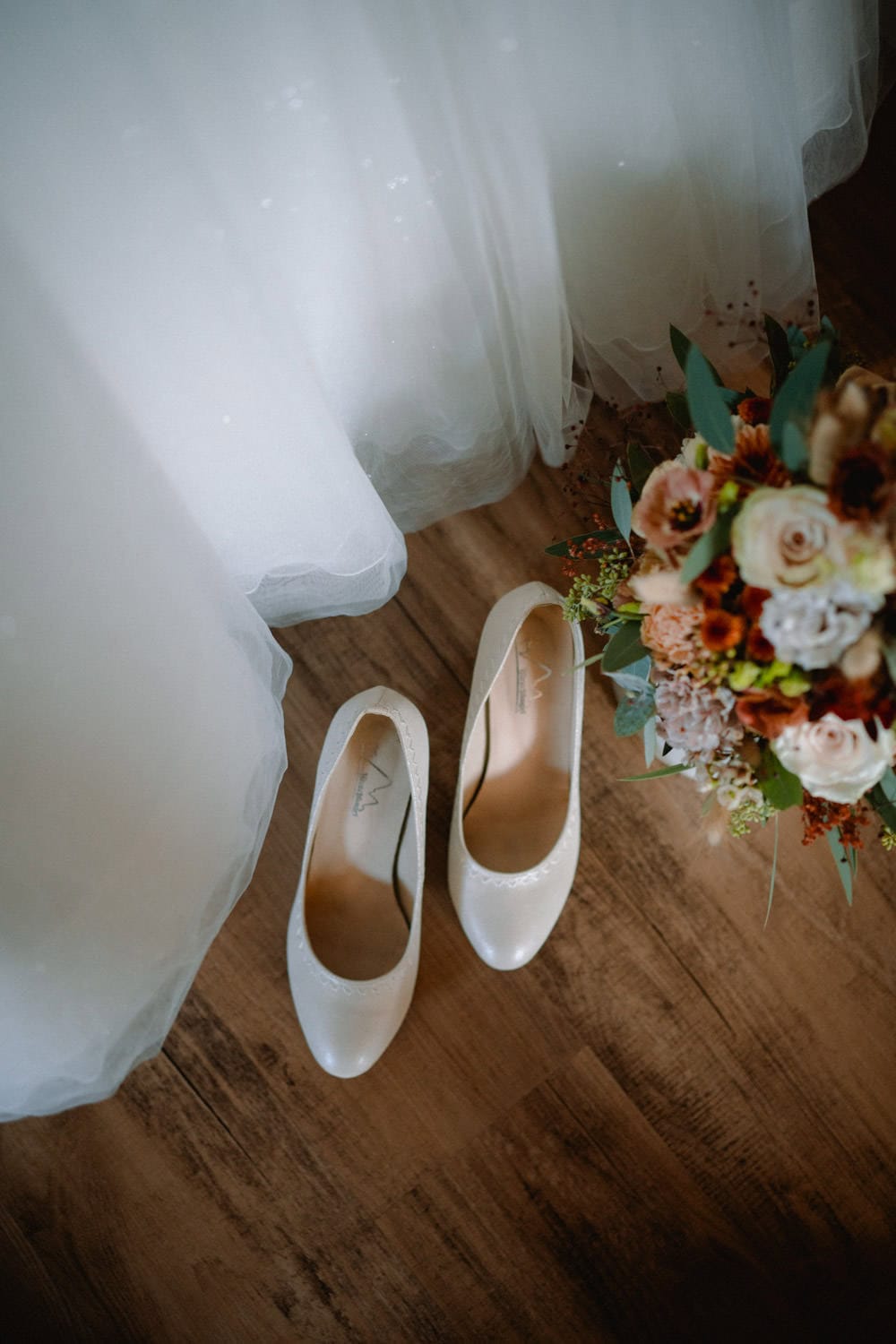 Ein Paar elegante weiße Schuhe ruht auf dem Holzboden von Hammerschloss Stodl Schmidmühlen, begleitet von einem Strauß Rosen und Grünpflanzen. Über den Schuhen ist teilweise ein fließendes weißes Kleid sichtbar, was den zeitlosen Charme der Szene unterstreicht.