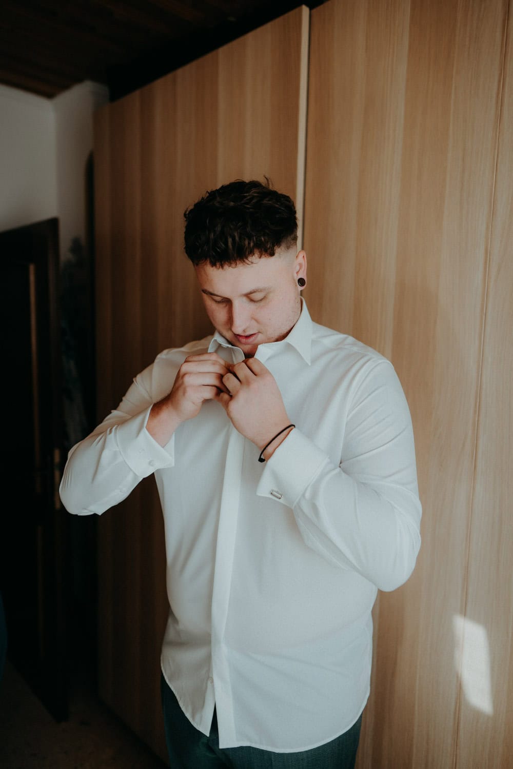 Eine Person mit kurzen Haaren und weißem Hemd steht vor einem Holzschrank im Hammerschloss Stodl und knöpft mit konzentrierter Miene ihr Hemd zu. Der Raum ist sanft beleuchtet, was eine warme Atmosphäre schafft.