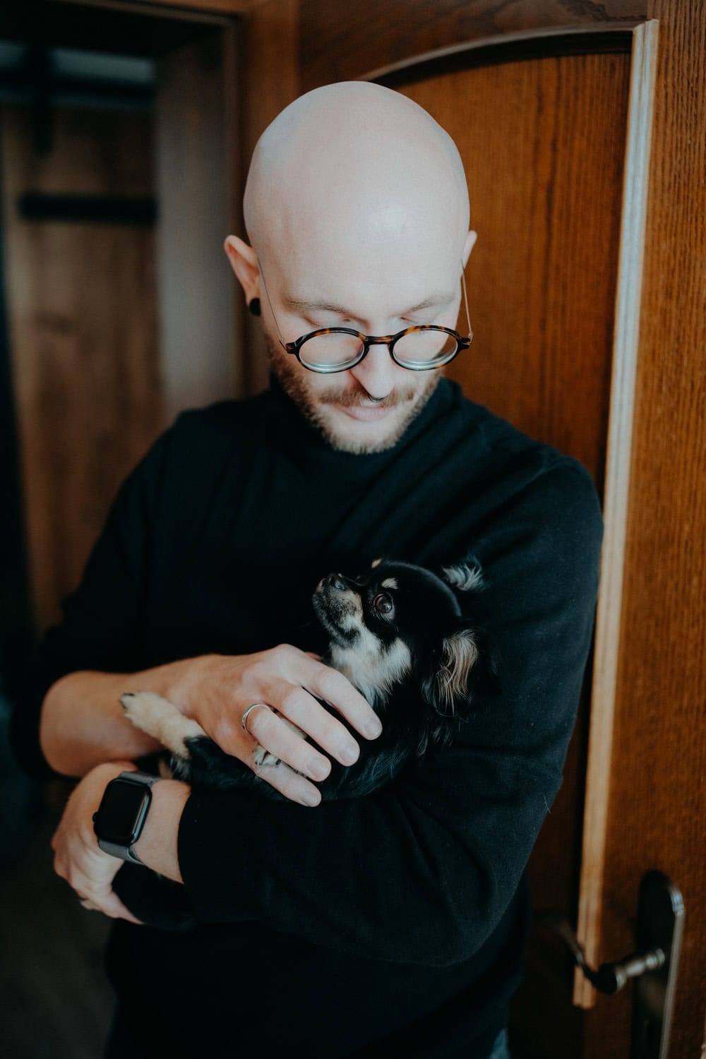 Vor einer Holztür hält eine kahl geschorene Person mit Brille und schwarzem Pullover einen kleinen schwarz-weißen Hund in ihren Armen und blickt ihn an. Dies erinnert an einen ruhigen Moment im Hammerschloss Stodl Schmidmühlen.
