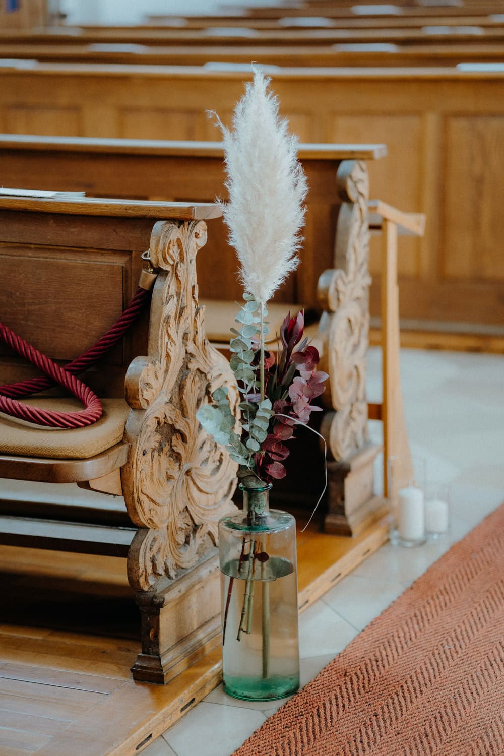 Eine große grüne Glasvase mit Pampasgras und Eukalyptuszweigen steht auf dem Boden neben einer kunstvoll geschnitzten hölzernen Kirchenbank, die denen im Hammerschloss Stodl in Schmidmühlen ähnelt. Die Bank ist mit rotem Seil und aufwendigen Mustern geschmückt, während im Hintergrund zwei Kerzen leise flackern.