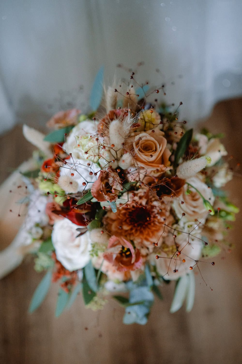 Ein rustikaler Brautstrauß besteht aus einer Mischung aus zartrosa Rosen, roten und weißen Blüten, akzentuiert mit grünen Zweigen und strukturierten Elementen. Auf einer Holzoberfläche im Hammerschloss Stodl Schmidmühlen platziert, erzeugt er einen natürlichen, eleganten Look, der perfekt zu einer Hochzeit auf dem Land passt.