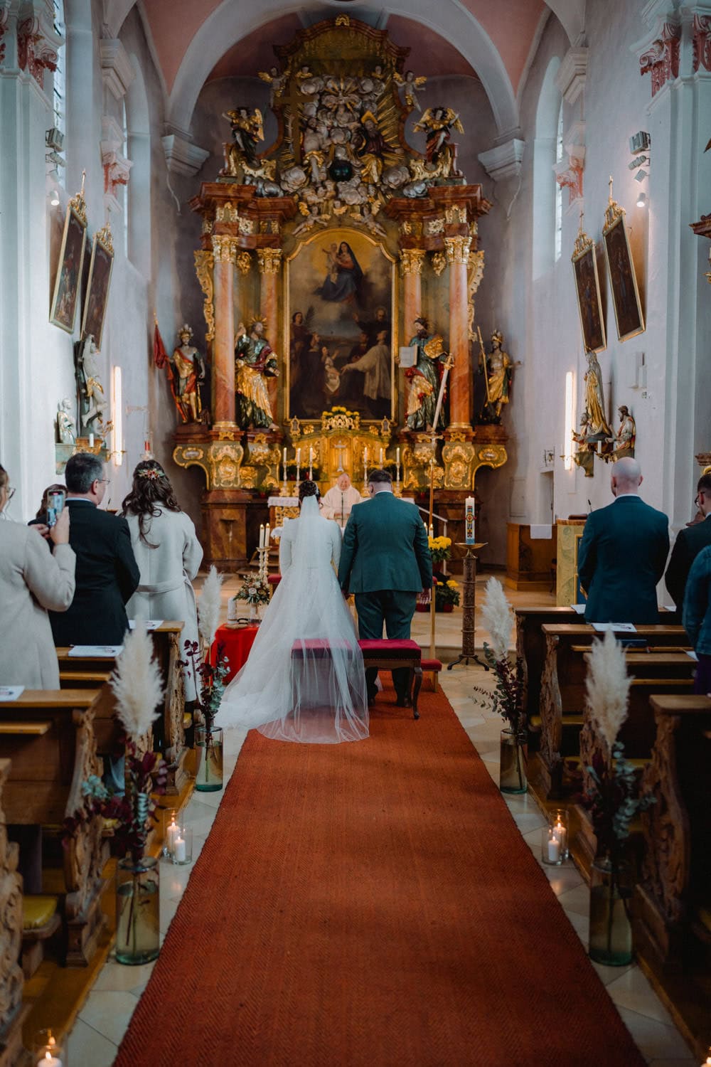 Ein Paar steht während einer Hochzeitszeremonie in der kunstvollen Hammerschloss-Stodl-Kirche am Altar. Die Braut trägt ein weißes Kleid und einen Schleier, der Bräutigam einen Anzug. Der Mittelgang ist mit Kerzen und dekorativen Pflanzen gesäumt, und auf beiden Seiten sitzen Gäste, was eine elegante Schmidmühlen-Atmosphäre schafft.