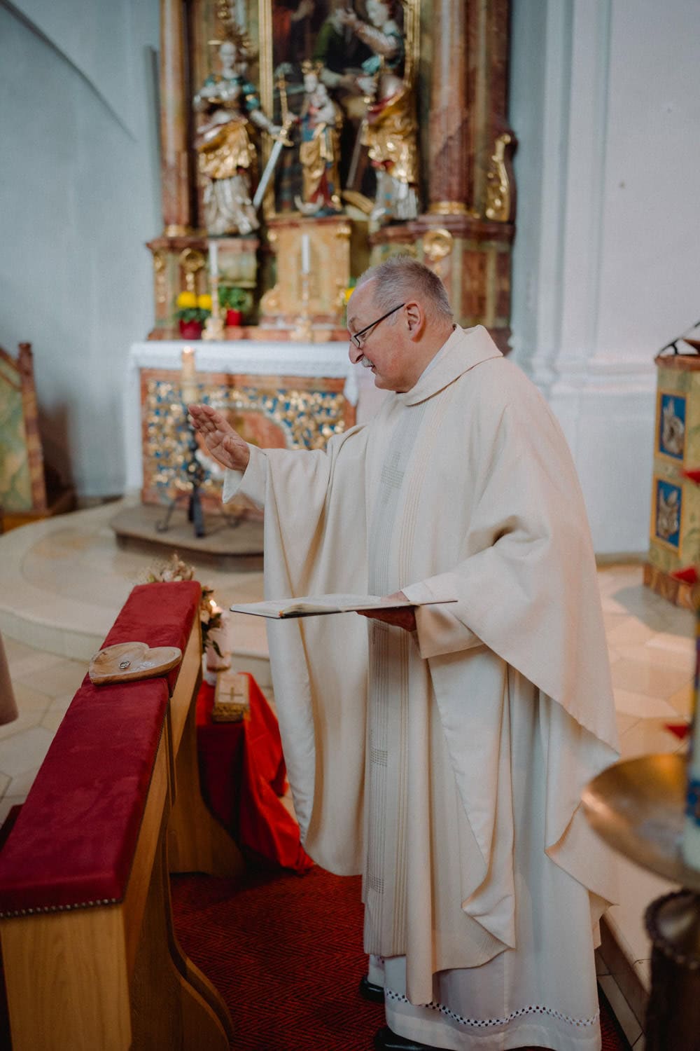 Ein Geistlicher in weißer Robe steht an einem Altar in der Kirche von Hammerschloss Stodl Schmidmühlen, hält ein Buch und macht mit einer Hand eine Geste. Der Hintergrund ist mit kunstvollen Verzierungen geschmückt.