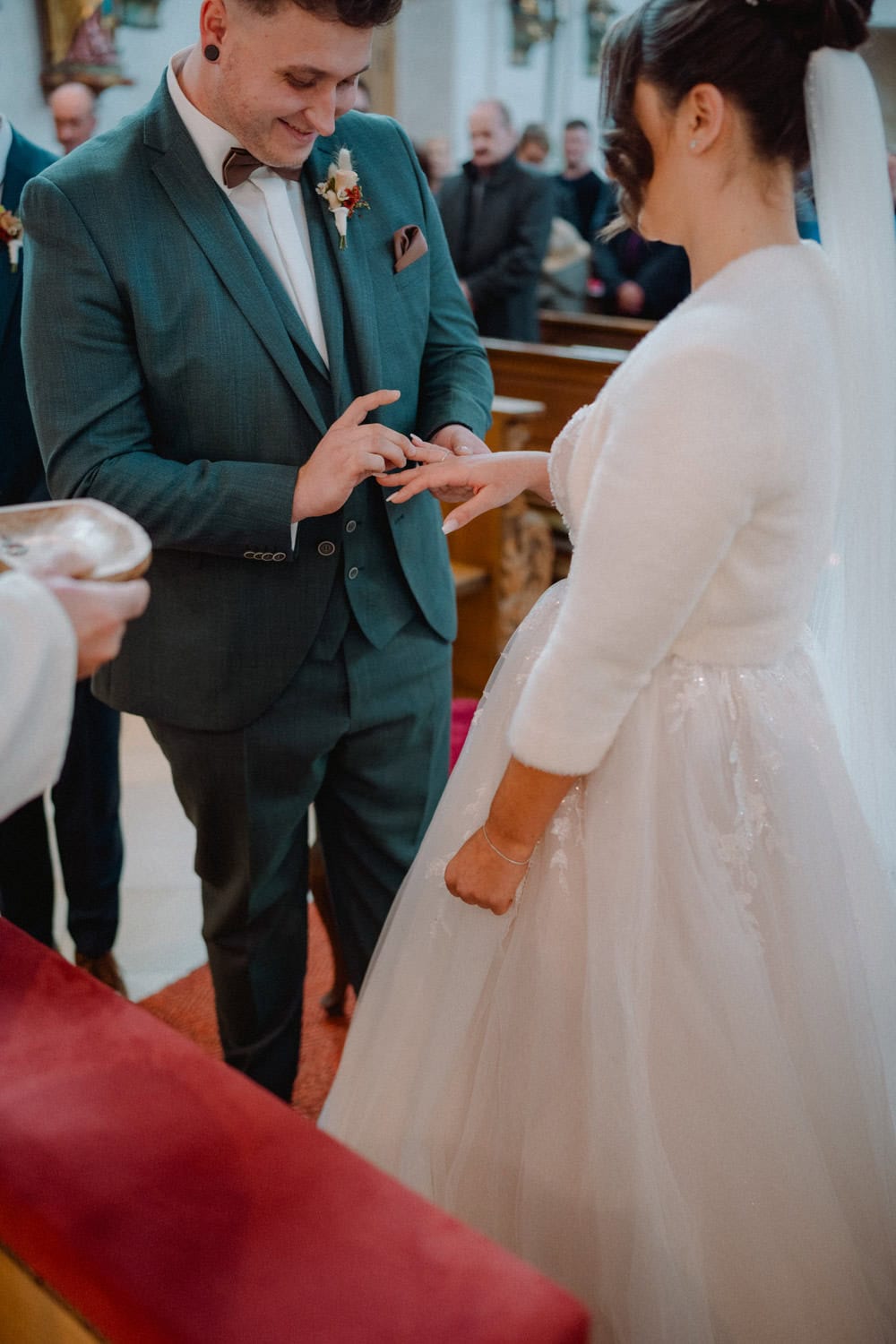 Ein Paar heiratet in der Kirche von Hammerschloss. Der Bräutigam, in einem blaugrünen Anzug, steckt der Braut einen Ring an den Finger. Die Braut, strahlend in einem weißen Kleid und Schleier, steht vor ihren Gästen, die voller Freude die wunderschöne Zeremonie verfolgen.