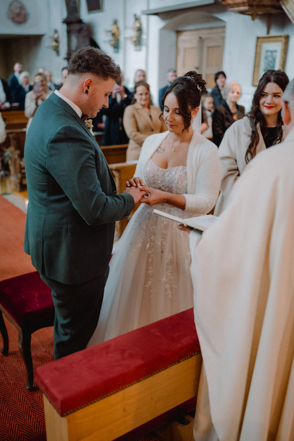 Während einer malerischen Zeremonie im historischen Hammerschloss Stodl in Schmidmühlen tauscht ein Paar Ringe aus. Die Braut in ihrem eleganten weißen Kleid und Schal steht neben dem Bräutigam im grünen Anzug, während die Gäste im Hintergrund diesen gefühlvollen Moment miterleben.