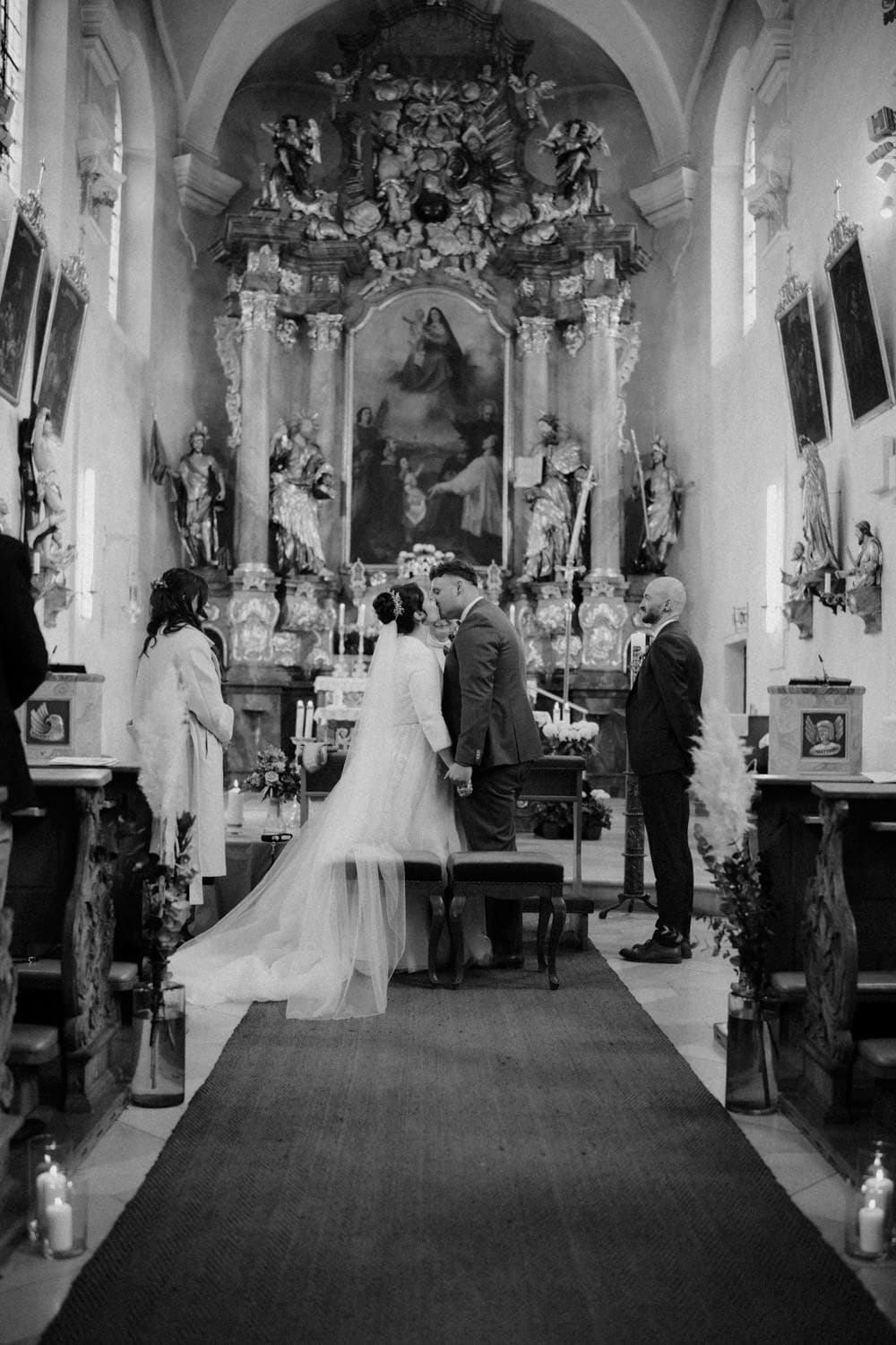 Braut und Bräutigam stehen vor einem Priester am Altar einer kunstvollen Kirche, die an Hammerschloss Stodl Schmidmühlen erinnert, umgeben von Gästen. Die Kirche zeichnet sich durch hohe Decken, religiöse Kunstwerke und aufwendige Dekorationen aus. Kerzen säumen den Mittelgang und tragen zur feierlichen Atmosphäre bei.