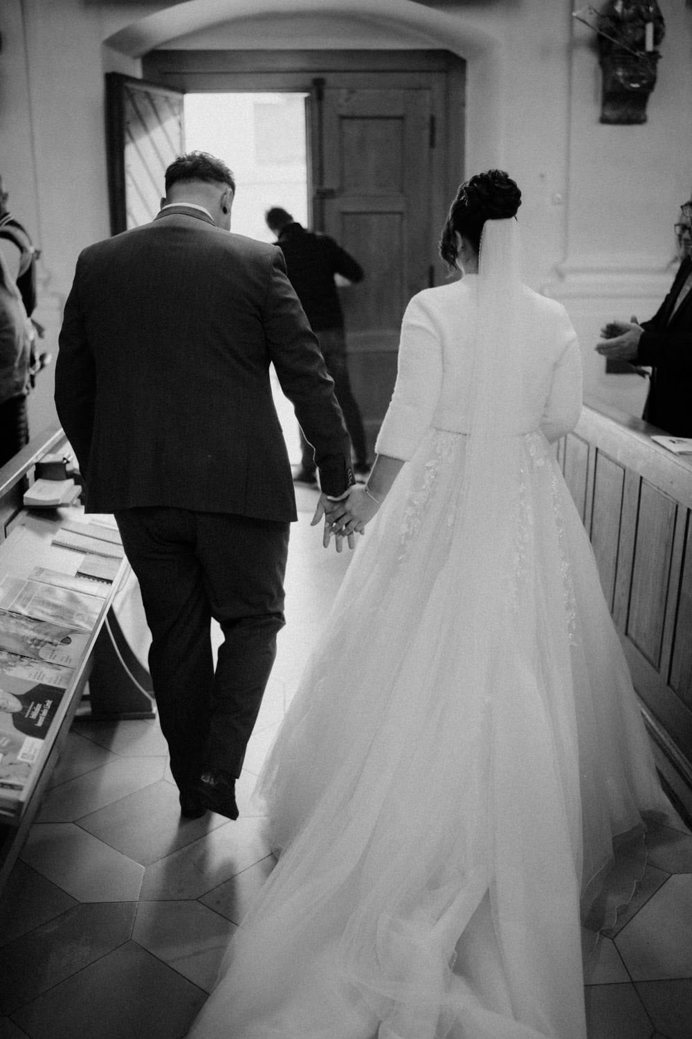 Ein Schwarzweißfoto zeigt Braut und Bräutigam, die Händchen haltend vor Hammerschloss Stodl Schmidmühlen von der Kamera weggehen. Die Braut trägt ein langes Kleid mit Schleier, während der Bräutigam einen Anzug trägt. Sie werden vom Licht eingerahmt, das durch eine offene Kirchentür vor ihnen fällt.